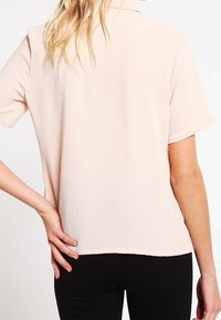 Kurzärmlige, hellrosa Bluse aus strukturiertem Stoff mit Kragen, von hinten gezeigt, kombiniert mit schwarzen Hosen.