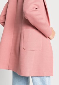 Cappotto rosa a motivo a spina di pesce con una singola tasca frontale, caratterizzato da una vestibilità rilassata e una consistenza morbida, abbinato a jeans azzurri chiari.