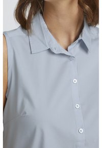 Błękitna bluzka bez rękawów z klasycznym kołnierzykiem, zapinana na guziki z przodu, o gładkiej fakturze. Zauważalne białe guziki i starannie wykonane szwy.