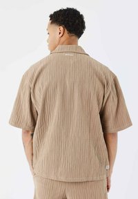 Camisa de manga corta texturizada en beige con un sutil patrón de costillas verticales. Cuenta con un cuello camp, así como una pequeña etiqueta de logo en el dobladillo.