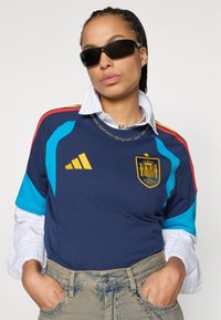 Μπλε και ναυτικό αθλητικό φανελάκι με κόκκινες και τιρκουάζ λεπτομέρειες, με κίτρινο λογότυπο και ρίγες. Φορεμένο πάνω από πουκάμισο με γιακά, σε συνδυασμό με γυαλιά ηλίου.