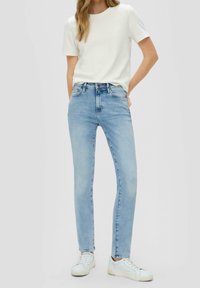 Femme portant un jean skinny bleu clair, un t-shirt blanc à manches courtes et des baskets blanches, debout avec une main dans la poche.