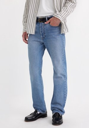 Personne portant un jean droit bleu clair, ceinture noire, mocassins en cuir noir, chemise blanche et chemise rayée boutonnée, une main dans la poche.