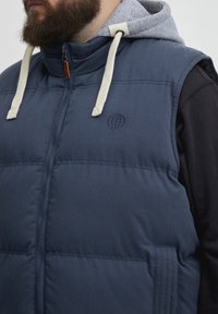 Mornarsko modra brezrokavnik z visokim ovratnikom, ki ima sivo kapuco in bež vrvico. Vključuje subtilno embosirano logo na prsih.