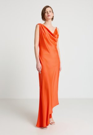 Femme debout dans une robe asymétrique sans manches orange avec un décolleté drapé et des talons blancs ouverts sur un fond clair uni.