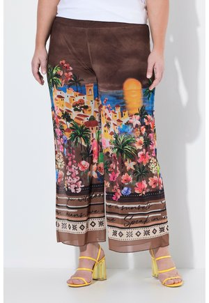 Pantaloni marroni a gamba larga con stampa colorata di tramonto, villaggio sul mare, fiori e palma, indossati con sandali gialli con tacco.