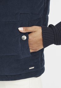 Marinblå manchesterväst med ficka med tryckknapp, ribbade mudd detaljer och en liten metallogga nära fållen. Texturerat tyg med rektangulära mönster.