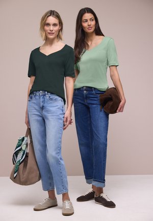 To kvinder iført afslappede V-hals bluser og jeans, den ene holder en taske med et tørklæde, den anden holder en brun jakke.