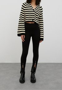 Pruhovaný zkrácený cardigan v černé a krémové barvě se zapínáním na knoflíky; spárovaný s černými ribbed legínami se zipy na kotnících a černými bojovými botami.