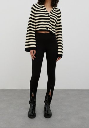 Gestreepte cropped cardigan in zwart en crème, met knoopsluiting; gecombineerd met zwarte ribgebreide leggings met ritsen bij de enkels en zwarte legerboots.