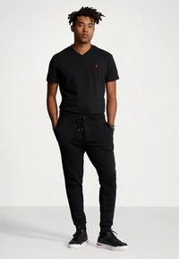 Črna bombažna majica z V-izrezom, kombinirana s črnimi trenirkami z elastičnim pasom in vrvico za zavezovanje. Oblečene s črnimi supergami.