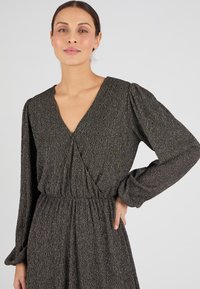 Abito testurizzato nero e oro con scollatura a V profonda, maniche lunghe a sbuffo e vita definita, con leggere rifiniture metalliche su tutto il vestito.