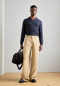 Suéter de punto azul marino con cuello tipo camisa, pantalones beige de pierna ancha y bolsa de viaje negra. Texturas suaves y líneas limpias en todo.