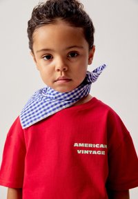 Jeune enfant aux cheveux bouclés attachés en arrière, portant un T-shirt rouge avec le texte « AMERICAN VINTAGE » et une écharpe à carreaux bleue autour du cou.