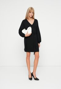 Mujer vestida con un vestido negro de manga larga y escote en V, con un lazo en la cintura, sosteniendo un clutch blanco y usando tacones altos negros, frente a un fondo blanco.