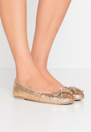 Pantoletas de ballet doradas brillantes con suela plana, punta redondeada y detalle decorativo de lazo de cuerda en la parte frontal. Textura suave con brillantes lentejuelas.