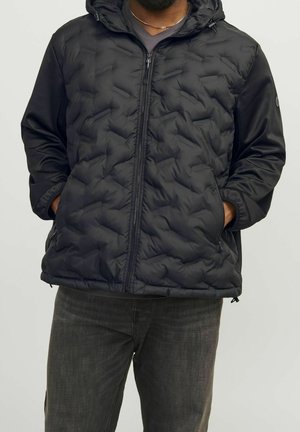 Mann trägt eine schwarze gesteppte Jacke mit Kapuze und Reißverschlusstaschen, steht mit den Händen in den Taschen vor einem schlichten Hintergrund.