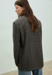 Blazer oversized marron à carreaux avec un motif écossais, épaules structurées et deux boutons aux poignets. Matériau en laine texturée.
