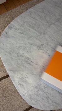 Okrągły biały marmurowy stół z szarymi żyłkami na beżowym, wykładanym dywanem podłodze, częściowo ukazujący biało-pomarańczowe prostokątne pudełko.