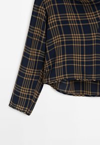 Chemise bleu marine à manches longues avec motif à carreaux beige et ourlet légèrement incurvé sur fond blanc.