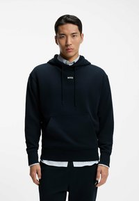 Young man wearing a black hoodie with "BOSS" text, layered over a white shirt, standing against a plain white background.