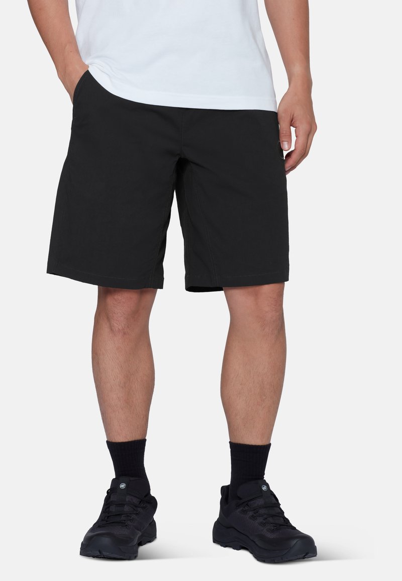 Mann trägt schwarze knielange Shorts, schwarze Socken, schwarze Sneakers und ein weißes Hemd, steht mit den Händen in den Taschen.