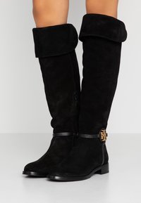 Botas de ante negras hasta la rodilla con un puño doblado, cierre lateral con cremallera y un acento decorativo en tono dorado en la correa del tobillo. Suela plana.