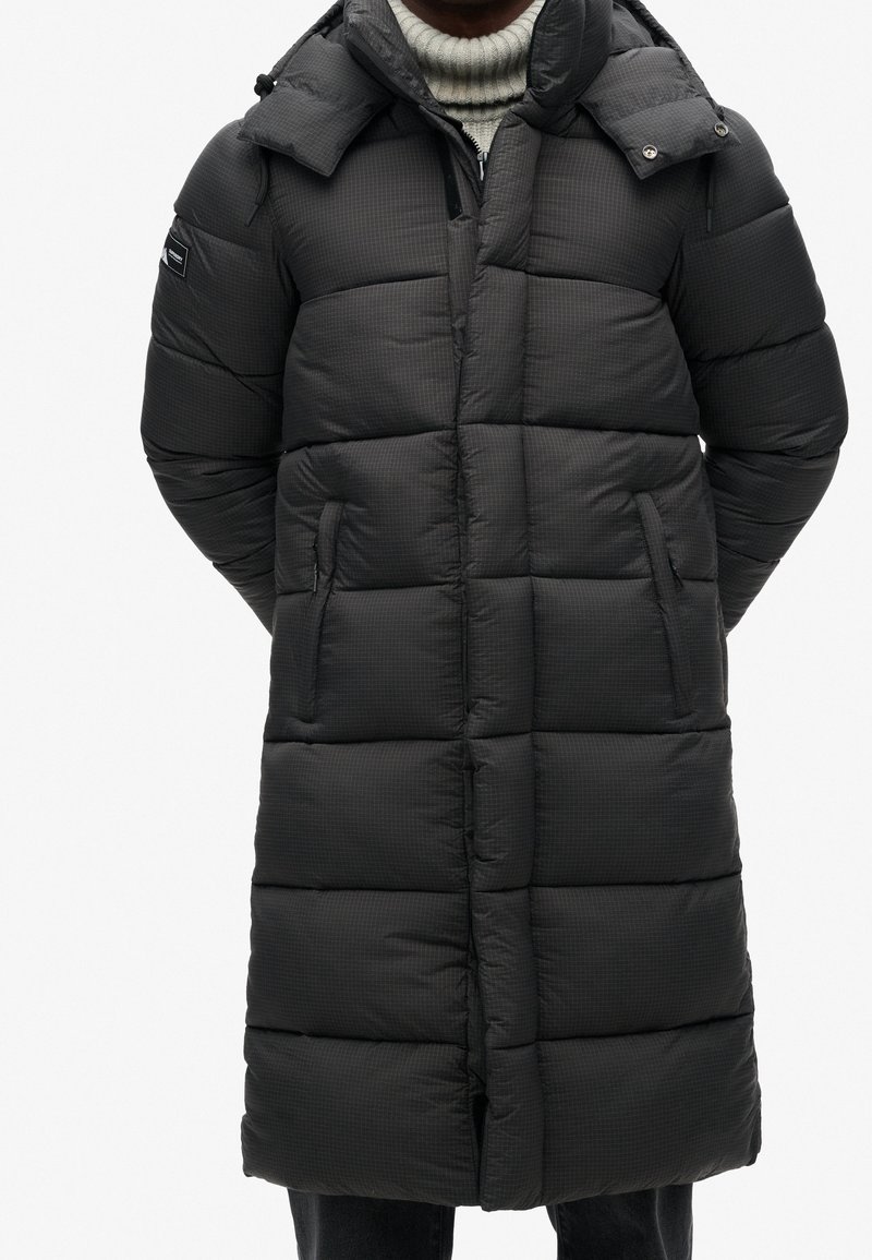 Długa, ocieplana kurtka w ciemnoszarym kolorze o kratkowanej fakturze, z kapturem, stojącym kołnierzem i bocznymi otworami na kieszenie. Zapięcie na zamek z przodu.