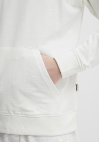 Fehér pamut pulóver elülső zsebbel, bordázott mandzsettákkal és hem. Az oldalán logócímke található. Puha tapintású és laza dizájn.