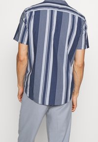Camicia a maniche corte di colore blu navy con strisce verticali bianche. Presenta un colletto con bottoni e un orlo curvo. Realizzata in un tessuto di cotone leggero.