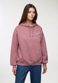 Felpa con cappuccio rosa realizzata in materiale morbido, con tasca frontale, cordini e ricami bianchi. Abbinata a jeans blu.