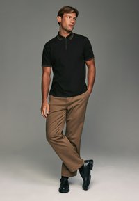Polo noir avec un col zippé, associé à un pantalon droit marron et des chaussures noires à enfiler. Tissu lisse avec des détails contrastés.