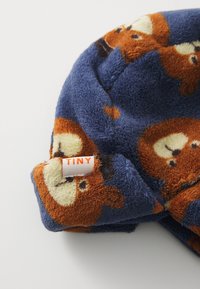 Blue fleece hat featuring an orange bear pattern. Includes a white tag labeled "TINY." Soft texture; rounded shape with a snug fit.