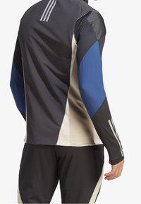 Camiseta deportiva de manga larga con paneles en negro, azul y crema, fabricada con un tejido texturizado y con tres franjas paralelas en la parte superior de la espalda.