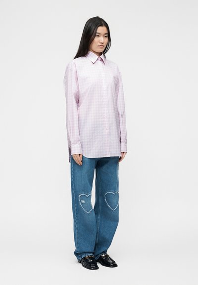 Γυναίκα με μακριά μαύρα μαλλιά που φορά ένα φαρδύ ανοιχτό ροζ καρό πουκάμισο, μπλε τζιν με μπαλώματα σε σχήμα καρδιάς στα γόνατα και μαύρα μοκασίνια.