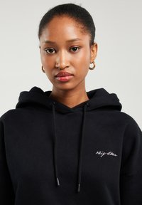 Noor naine, kes kannab kuldseid rõngakujulisi kõrvarõngaid ja musta kapuutsi, mille rinnal on tikitud "Big Star", vaatab kaamerasse neutraalse ilmega.