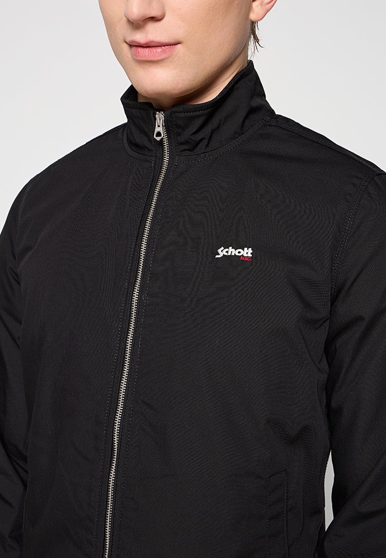 Gros plan sur une personne portant une veste zippée noire avec un col haut et le logo "Schott NYC" sur la poitrine.