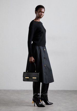 Schwarzer, gerippter Pullover, knielanger schwarzer Rock mit Knopfdetails, kombiniert mit schwarzen Stilettos mit goldenen Absatzdetails, während eine schwarze Handtasche getragen wird.