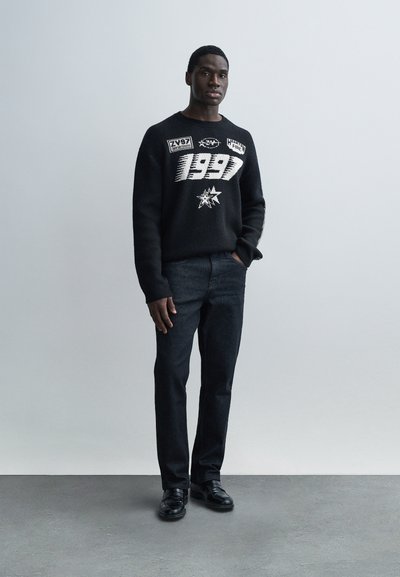 Černý pletený svetr s bílými grafickými motivy a textem. Kombinován s tmavými džínami a černými botami. Současný přiléhavý střih.