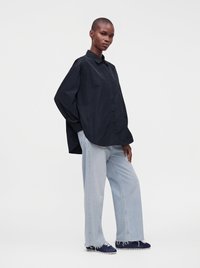 Personne portant une chemise noire ample boutonnée, un jean large bleu clair et des baskets bleu foncé, debout devant un fond blanc uni.