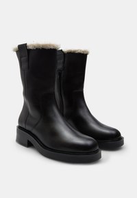 Botas de cuero negras hasta el tobillo con forro de pelaje sintético, cierre lateral y tacón cuadrado. Textura suave con un diseño elegante y minimalista.