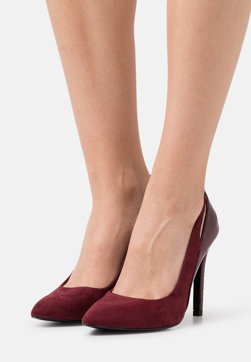 Paire de jambes portant des escarpins en daim rouge foncé à talons hauts et bouts pointus sur un fond clair uni.