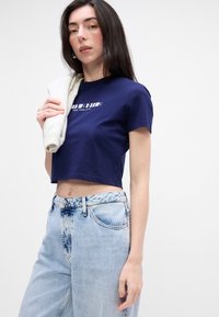 Tamsiai mėlynos spalvos trumpa marškinė su baltais logotipo užrašais, derinama su šviesiai mėlynomis aukštu juosmeniu džinsais, turinčiais nudilimą ir kišenių detales.