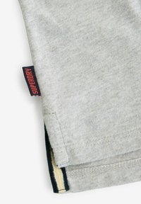Grå bomullströja med fåll som har en röd och marinblå etikett med "SUPERDRY." Dubbel söm med en gul accent vid sidospåret.