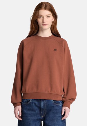 Ung kvinde med langt rødt hår iført en brun sweatshirt og blå jeans, står foran en ensfarvet lys baggrund.