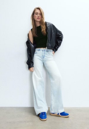 Kobieta w czarnej skórzanej kurtce zsuniętej z jednego ramienia, czarnym krótkim topie, szerokich jasnoniebieskich jeansach i niebieskich trampkach, stojąca przy białej ścianie.