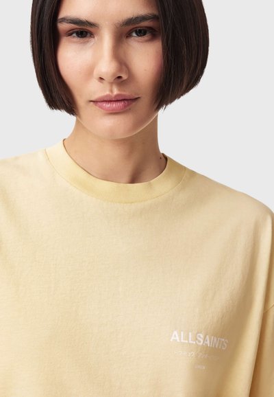Jeune personne aux cheveux courts et foncés portant un t-shirt AllSaints jaune pâle avec un lettrage blanc sur un fond clair uni.