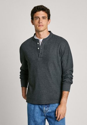 Camisa henley gris de manga larga con un acabado texturizado, tapeta de tres botones y cuello redondeado, combinada con jeans de mezclilla azul.