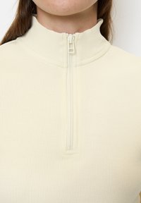Helles cremefarbene, gerippte Zip-Up-Oberteil mit Stehkragen und halblangem Reißverschluss. Glatte Textur mit sichtbar genähten Details.