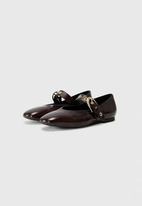 Ballerines en cuir marron avec un nez pointu, une seule sangle, un accent en métal doré et une semelle plate. Texture lisse avec des détails de design minimaux.